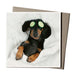 A greeting card featuring an illustration of a dog with cucumber slices on its eyes, lying on a white surface, with a brown envelope included.